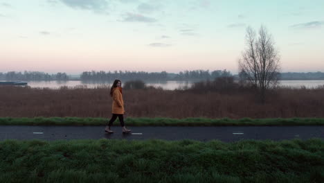 Paralex-shot,-girl-walking-down-on-a-dam,-at-early-morning-with-the-river-in-the-background
