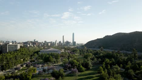 Rückwärtige-Luftaufnahme-Des-Zweihundertjährigen-Parks-Von-Vitacura-In-Der-Stadt-Santiago-De-Chile-Mit-Dem-Finanzviertel-Am-Horizont---Drohnenaufnahme