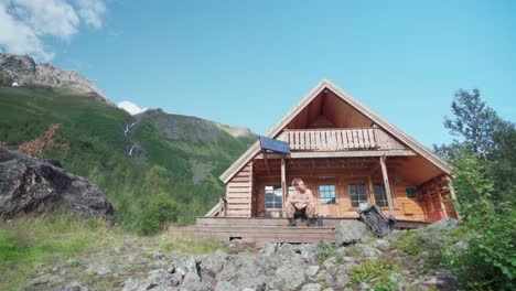 Shirtless-Man-Tourist-Relaxing-Sitting-On-The-Front-Porch-Steps-Of-A-Wooden-Cabin-In-Lyngsdalen-Valley,-Norway