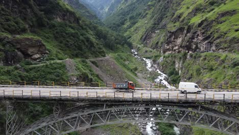 A-drone-shot-of-commercial-vehicles-traveling-on-a-suspended-bridge-on-a-river-valley-in-the-remote-highway-of-Himachal-Pradesh,-at-the-foothills-of-the-Indian-Himalayas