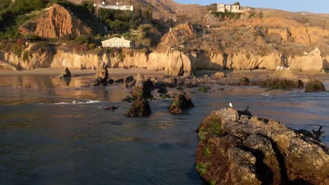 Aerial-view-of-rocks-by-the-shore-of-El-Matador-Beach-in-Malibu-California,-birds-standing-on-top-of-the-rocks-and-waves-rolling-on-the-shore-during-golden-hour,-gorgeous-landscape