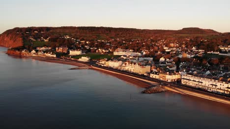 Aerial-Above-English-Channel-With-View-Of-Sidmouth-Town-Esplanade-Bathed-In-Golden-Sunrise-Light