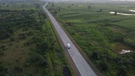 Drone-following-a-goods-carrier-van-traveling-on-a-rural-Indian-road-of-good-quality-with-the-surroundings-of-green-farmlands-and-village-homes