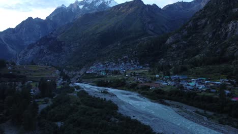 Una-Foto-De-Un-Dron-De-Un-Pequeño-Y-Pintoresco-Pueblo-Remoto-A-Orillas-Del-Río-Baspa-Con-Los-Picos-Nevados-Del-Himalaya-Indio-Al-Fondo