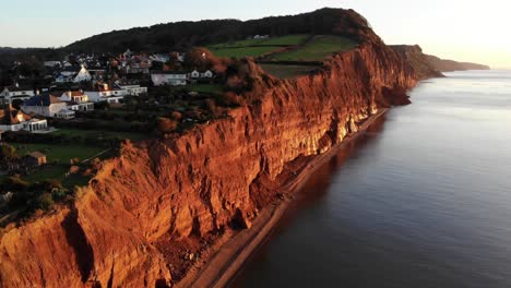 Aerial-forward-shot-of-the-red-Jurassic-cliffs-at-Sidmouth-South-West-England-at-sunrise-bathed-in-golden-sunlight