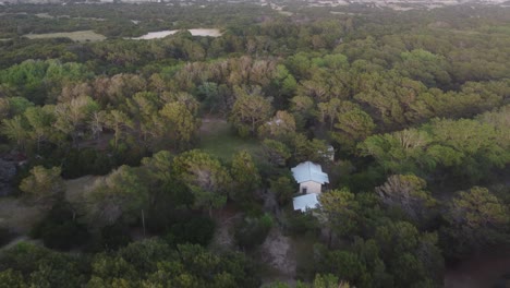 Aerial-moving-shot-of-only-white-house-in-middle-of-the-beautiful-forest-on-a-sunny-day-out-at-Mar-de-las-Pampas,-South-America