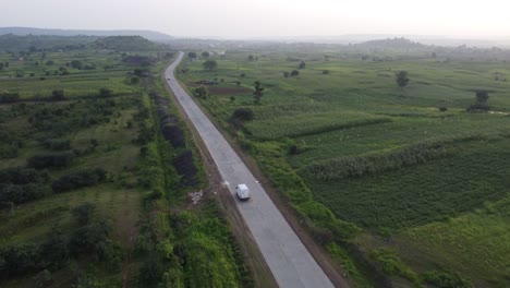 Drone-following-a-goods-carrier-van-traveling-on-a-rural-Indian-road-of-good-quality-with-the-surroundings-of-green-farmlands-and-village-homes