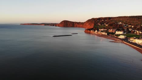 Aerial-Along-Sidmouth-Coastline-With-Offshore-Breakwaters-Bathed-In-Golden-Sunrise-Light