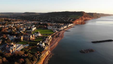 Aerial-Along-Coastline-With-Sidmouth-Town-Bathed-In-Golden-Sunrise-Light