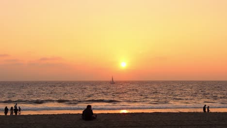Wunderschöner-Sonnenuntergang-Am-Strand-Mit-Einem-Boot,-Das-Auf-Dem-Meer-Segelt,-Und-Menschen,-Die-Auf-Dem-Sand-Spazieren-Gehen-Und-Die-Wellen-Betrachten