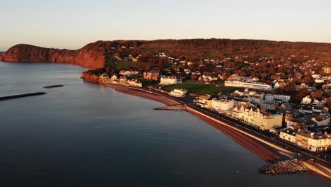 Aufsteigende-Luftaufnahme-Der-Stadt-Sidmouth-Und-Der-Juraküste-Südwestenglands,-Die-Die-Goldene-Rote-Farbe-Des-Schönen-Morgensonnenaufgangs-Widerspiegelt
