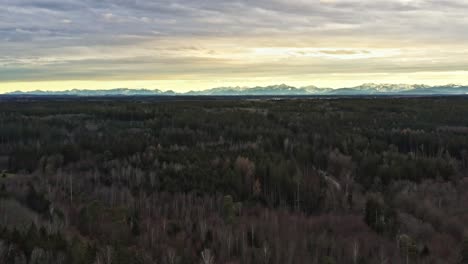 In-mountain-range---drone-flight-backward-over-a-wide-forest-with-sun-behind-clouds