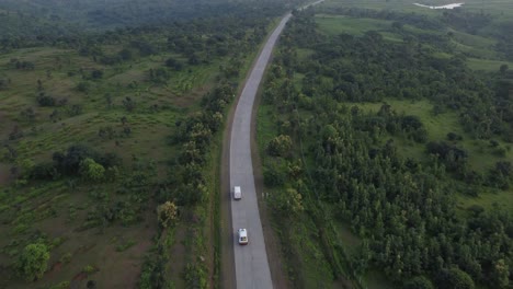 Drone-following-an-ambulance-overtaking-a-goods-carrier-van-traveling-on-a-remote-rural-Indian-road-of-good-quality-with-the-surroundings-of-natural-forest-and-green-farmlands-and-village-homes