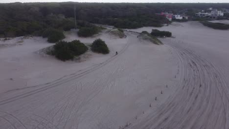 Aerial-view-of-a-person-driving-a-motorbike-on-the-beach-sand-in-a-forest-nature-background-in-Mar-de-las-Pampas,-South-America
