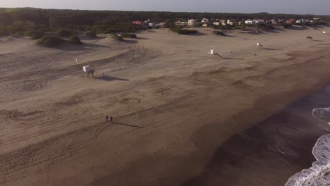 Slow-motion-aerial-drone-shot-of-an-two-people-walking-on-the-sand-in-front-of-the-beach-water-waves-on-a-cloudy-sunset-day-at-Mar-de-las-pampas,-Argentina