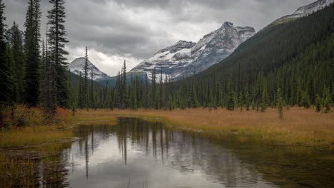 Time-lapse,-cold-autumn-day-in-mountains-of-Canada,-dramatic-dark-clouds-above-snow-capped-summits-and-valley-with-conifer-forest-and-pond-water