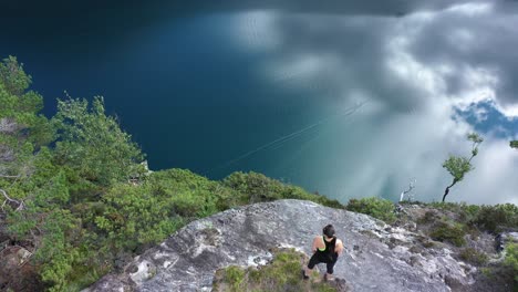 Flying-overhead-of-woman-standing-on-cliffs-edge---Spectacular-and-dangerous-cliff-Slottet-in-Modalen-Norway---Woman-enjoying-viewpoint-and-fjord-with-sky-reflections
