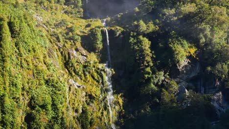 Tilt-down-shot-of-epic-cascading-waterfall-between-green-mountain-walls-lighting-by-sun---Fiordland-National-park,New-zealand