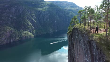 Woman-enjoying-spectacular-view-from-edge-of-tall-mountain-cliff-Slottet-in-Modalen-Norway---Forward-moving-aerial-close-to-cliff-in-stunning-fjord-landscape-with-boat-passing-on-fjord-below---Norway