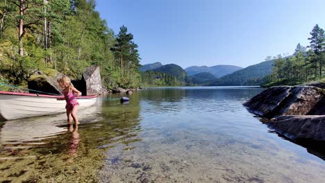 Young-blonde-child-in-pink-swimsuit-wading-and-playing-with-boat-at-beach---Cozy-bay-in-freshwater-lake-during-summer-in-Norway---Western-Norway-static