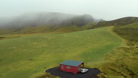 Aerial-icelandic-Landscape,-lonely-red-cabin-on-Mountain-green-fields