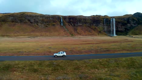 Tracking-shot-follow-car-passing-by-Skogafoss-waterfall,-Icelandic-Landscape,-Aerial