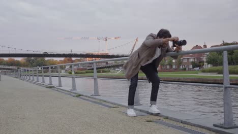 Stylish-young-man-with-a-professional-camera-trying-to-find-a-good-position-for-a-shot-on-a-street-along-a-river-in-an-urban-area