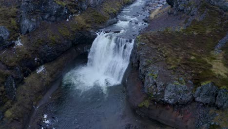 Ascending-top-down-shot-of-vast-waterfall-falling-into-Sela-River-during-cloudy-day-on-Iceland-Island,Europe---Spectacular-volcanic-scenery-with-natural-floating-melt-water