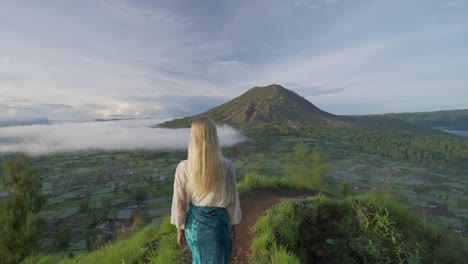 Woman-standing-at-Kayupadi-viewpoint-enjoying-view-of-Mount-Batur,-golden-hour-sunlight