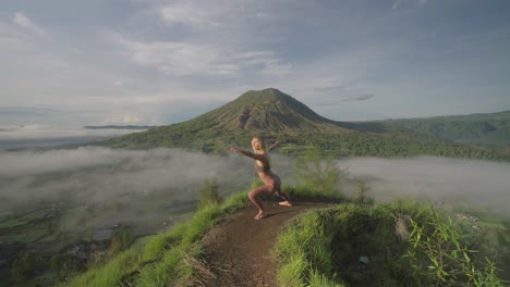 Fit-woman-in-Warrior-Pose-on-mountain-edge-overlooking-tropical-landscape,-morning-sunlight