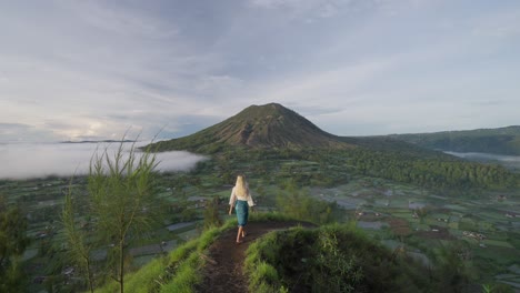Blonde-Frau-Mit-Traditionellem-Sarong-Am-Aussichtspunkt-Kayupadi,-Die-Hände-Zur-Begrüßung-Hebend,-Mount-Batur-Caldera