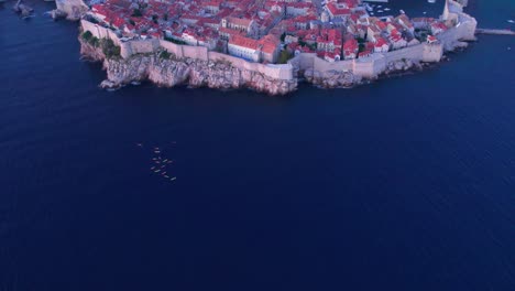 Group-of-kayakers-floating-close-to-rocky-shore-of-medieval-Dubrovnik,-aerial