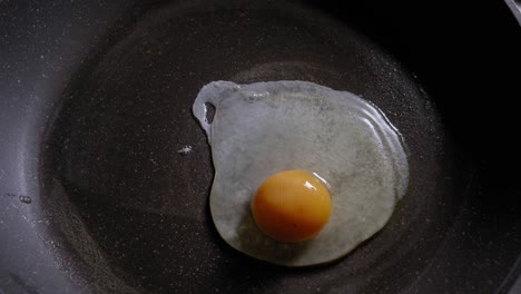 A-top-down-close-up-shot-of-a-fresh-egg-being-poured-onto-a-lightly-greased-frying-pan,-the-raw-egg-changes-colour-as-it-starts-to-cook-and-splutters-and-bubbles-on-the-surface-of-the-hot-pan