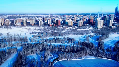 Luftaufnahme-Von-Edmonton-Downtown-West-Enthüllung-Der-Stadt-Downtown-Hochhäuser-Im-Hintergrund-Und-Im-Vordergrund-Der-River-Park-Schneebedeckte-Kiefern-öffentliche-Outdoor-Eisbahn-Langlauf