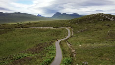 Male-on-longboard-riding-downhill-on-a-gray-asphalt-road-between-the-mountains-of-the-Scottish-Wilderness-on-a-cloudy-day