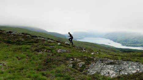 Männchen-In-Schwarzer-Trainingskleidung,-Das-über-Die-Grünen-Hügel-Mit-Lochbesen-Unter-Den-Tief-Hängenden-Wolken-Im-Hintergrund-In-Der-Nähe-Von-Ullapool,-Schottland,-Läuft