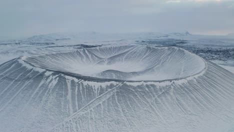 Aerial-backwards-shot-of-snowy-Hverfjall-Crater-Volcano-during-cloudy-day-in-Iceland
