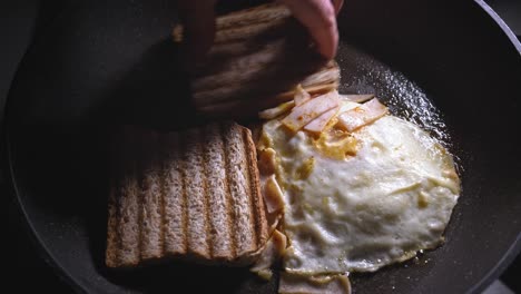A-close-up-shot-of-toasted-bread-being-placed-into-a-frying-pan-alongside-a-fried-egg-topped-with-ham-and-melted-cheese,-the-filling-is-placed-on-the-bread-to-complete-the-assembly-of-the-sandwich