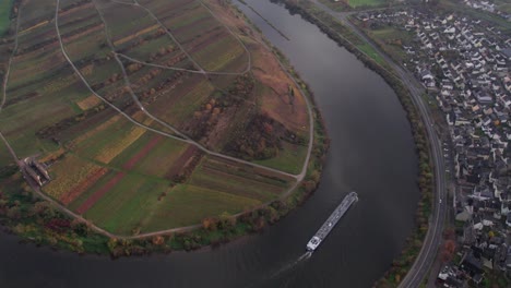 Industrial-cargo-ship-moving-through-Moselle-Bremm-river-bend-passing-vineyard,-aerial