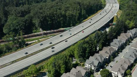 Luftdrohnenansicht-Eines-Zuges,-Der-Unter-Einer-Stark-Befahrenen-Autobahn-Im-Großraum-Vancouver-Fährt