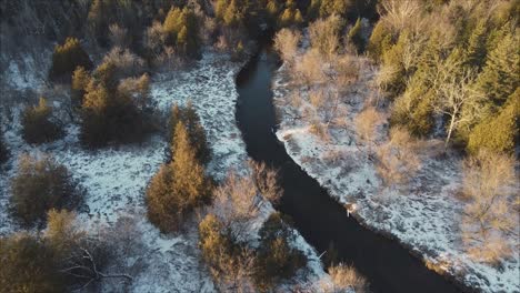 Aerial-Flyover-Of-River-In-A-Winter-Forest-Landscape,-Trees-Lit-At-Sunset