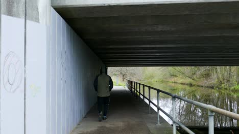 The-back-of-an-anonymous-man-walking-under-an-overhead-bridge,-the-footpath-running-parallel-to-the-bank-of-the-Thetford-Little-River-in-Norfolk,-England