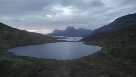 Aerial-drone-view-low-over-a-lake-towards-mountains,-sunrise-in-cloudy-Lofoten,-Norway