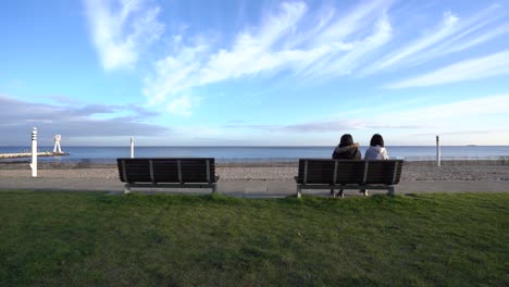 Scenic-view-of-girls-sitting-and-talking-on-a-bench-and-looking-at-the-beach,-wide-static