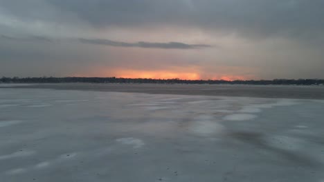 Luftaufnahme-Eines-Zugefrorenen-Sees-Mit-Kühlen-Mustern-Auf-Dem-Eis-Mit-Einem-Farbenfrohen-Sonnenuntergang-Am-Horizont