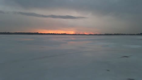 Aerial-view-of-a-frozen-lake-with-a-colorful-sunset-on-the-horizon-on-a-cloudy-day