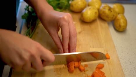 Female-Hands-Cutting-a-Carrot-on-a-Wooden-Cutting-Board-With-Parsley-and-Potatoes-in-the-Background