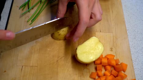 Weibliche-Hände-Schneiden-Kartoffeln-Auf-Einem-Holzschneidebrett-Mit-Petersilie-Und-Gewürfelten-Karotten-Im-Hintergrund