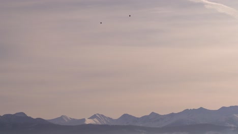 Two-hot-air-balloons-flying-high-above-snowy-mountain-landscape-at-sunset