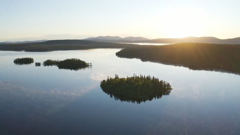 Aerial-view-high-above-a-blue-foggy-lake-during-sunrise-showing-small-islands-and-mountains-in-the-background-as-sun-is-giving-golden-hour-light-to-the-landscape-filmed-in-Lapland-Finland